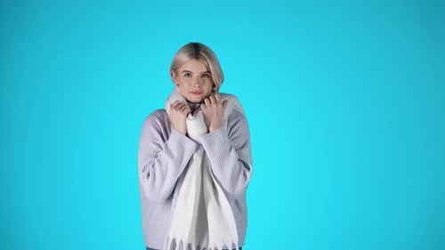 Portrait Of Young Blonde Woman Feeling Cold, Wearing Scarf and Grey Sweater, Studio Shot With Blue B