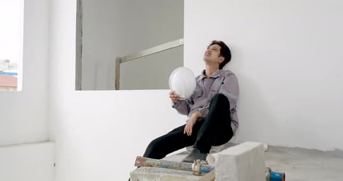 Asian male contractor sits on concrete ledge holding helmet while reviewing unfinished building