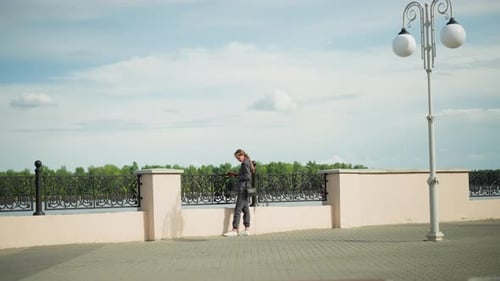 Lady in Grey Reading By Riverfront with Lamp Post and Trees in Distance