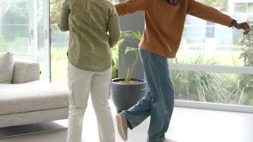Casal afro-americano praticando passos de dança, braços estendidos por portas de vidro deslizantes na sala de estar