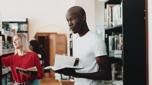 Young Africanamerican Student is Standing in Library with Book at Hands and Reading Side View