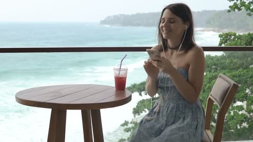 Happy Woman Listening to Music and Drinking Cocktail Sitting on Terrace 30s