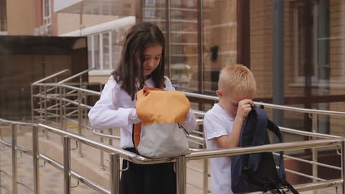 A Boy and a Girl in School Uniforms Look Into Open Backpacks Near the School