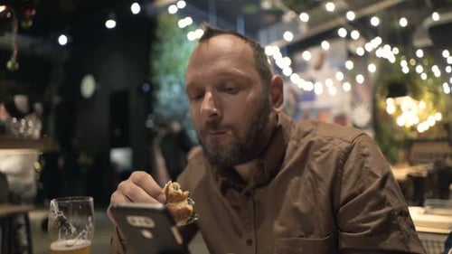 Young man enjoys burger and texts on smartphone in a sunny cafe