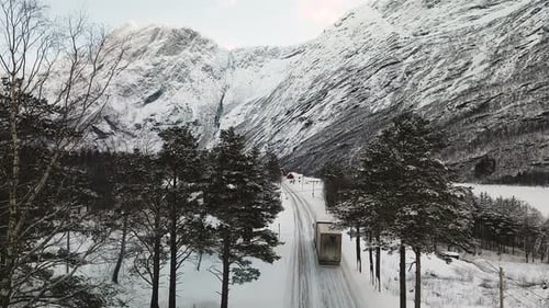 Aerial View Of Cargo Truck Driving On Snowy Asphalt Road In Canada In Winter With Rocky Mountains In