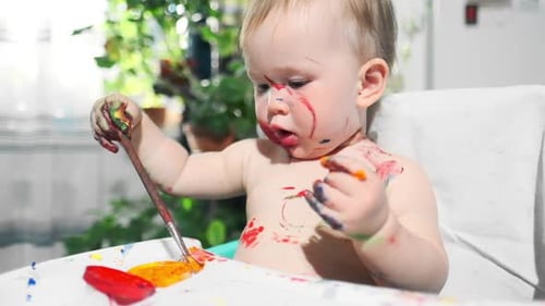 Baby paints at home. Portrait of the infant baby enthusiastically drawing with brushes and paints an