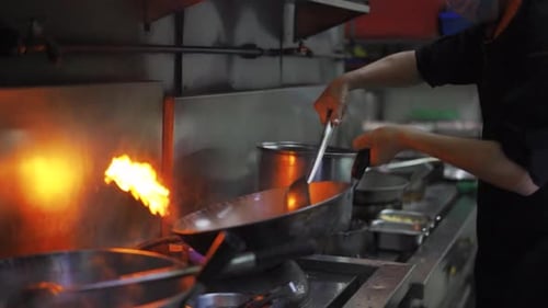 Chef Tossing Wok While Sauteing Food Over High Flame In Kitchen Of A Restaurant. - slow motion