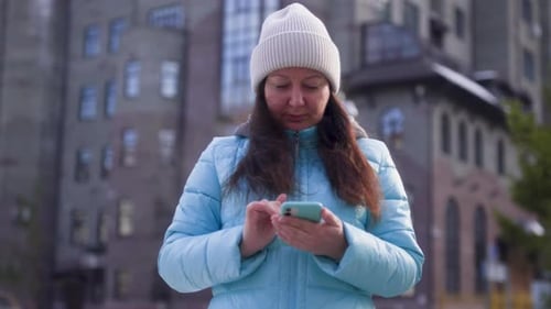 Middle Aged Brunette Woman Types Message in Smartphone on City Street in Winter