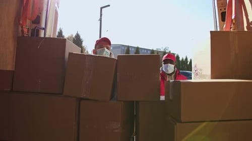 Afroamerikanische und kaukasische Kuriere mit Gesichtsmasken bringen Paketboxen von einem Van auf einen Trolley