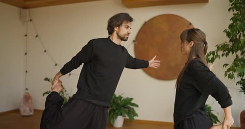 Two Adults Wearing Black Outfits Practice a Balance Pose Indoors Holding One Knee Up and Extending