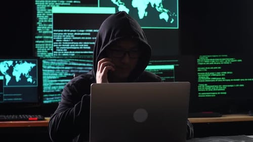 Hacker em um capuz falando em um smartphone enquanto trabalha em um laptop com mapas e dados em telas