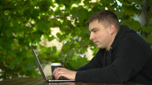 A Young Man at a Table in a Gazebo Works on a Laptop During the Day A Man in a Black Hoodie with a
