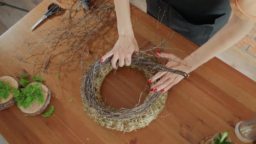 Female Florist Hands Arranging Twigs on Hay Wreath Base on Wooden Table