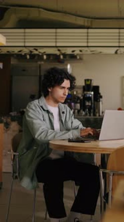 Focused Young Man Working on Laptop in Modern Casual Cafe Setting Concentrating on Remote Tasks