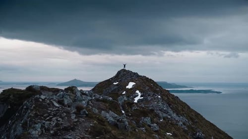 Person standing on top of a large mountain with their arms up in the air, surrounded by seascape in