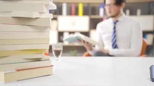 Man Reads Book at Office Desk with Stacked Books