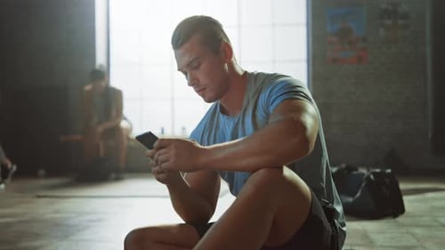 Handsome and Masculine Athletic Young Man is Using a Smartphone while Sitting on a Floor in a Loft