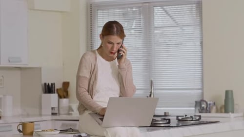 Woman on Phone Using Laptop in Kitchen