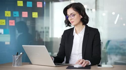 Confident business woman wearing glasses sitting at work desk with laptop in office. Professional