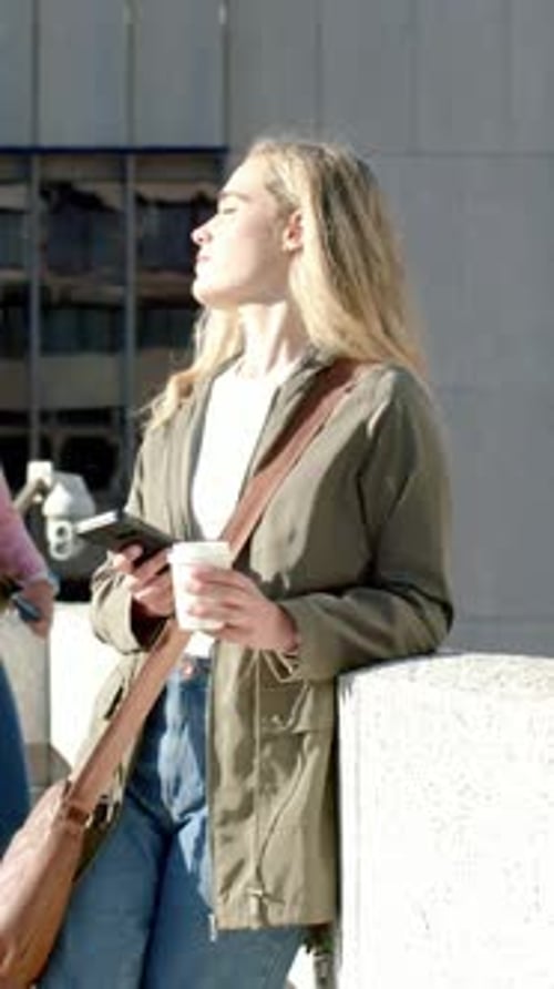 Vertical video: Tilting face toward sun, woman in olive jacket leaning on railing, with phone, cup