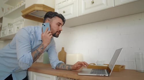 Adult Man Talking on Phone Using Laptop in Kitchen