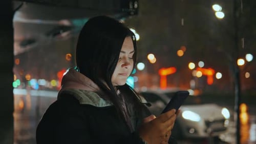 Ein Mädchen im Regen steht mit einem Telefon vor dem Hintergrund verschwommener Laternen