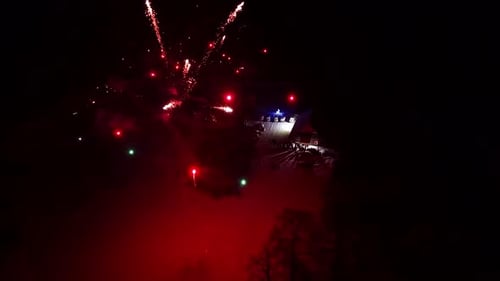 Night fireworks burst above a dark waterfront with scattered city lights, creating a festive