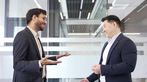 Two businessmen in formal suits shaking hands after deal while standing in a modern business office.