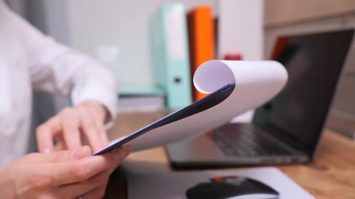 Person's Hands Holding and Reviewing a Document on a Clipboard at an Office Desk with a Laptop and