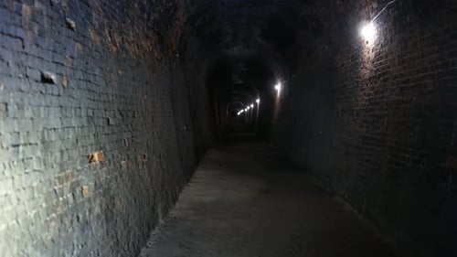 A long, brick-lined tunnel underground with a slightly curved ceiling is illuminated lights along th
