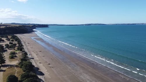 Aerial shot, flying over a beautiful beach with people on a sunny day in Long Bay, New Zealand.