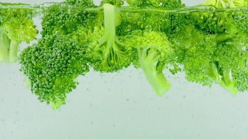 Green broccoli florets fall into boiling water with air bubbles.