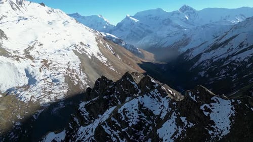 Aerial snowy winter mountain range ridgeline with steep valleys below on sunny day, flying around pe