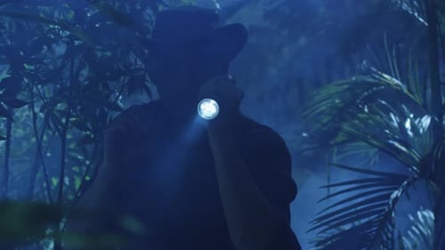 Adventurer in Hat Walking through Jungle Forest in Night. Using Flashlight