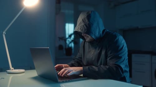 Vermummter Hacker tippt wütend und frustriert auf einem Laptop in einem dunklen Raum