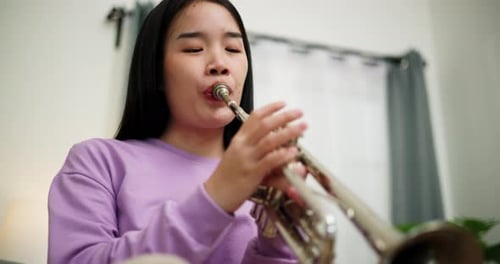 A young woman practices playing the trumpet while sitting on a sofa in a cozy living room.