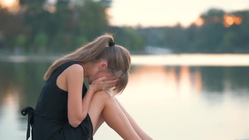 Upset Woman Sitting by Lake at Sunset