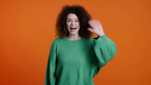 Cheerful Stunning Young Woman with Stylish Afro Hairstyle Waves Hello with Hand on Color Background