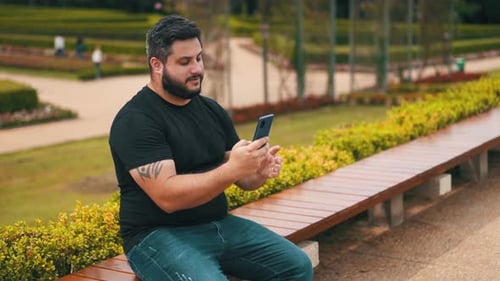 Young bearded man on jeans pants using his cell phone outdoors while seated on a bench