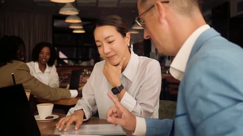 Asian woman and coworker talking about work while using laptop in modern coffee shop.