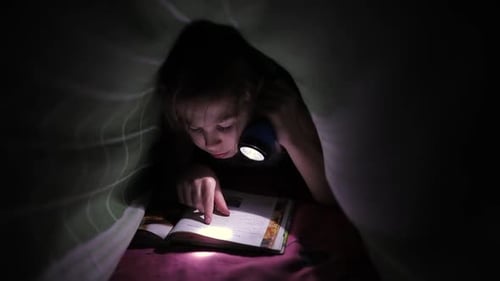 Little Child Girl Reading a Book with a Flashlight Covered with Blanket