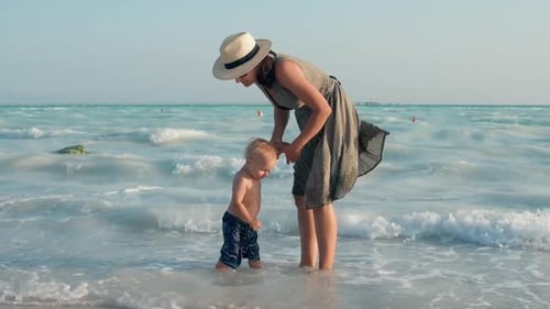 Kid Sitting In Sea Water At Family Weekend. Beautiful Woman Speaking To Little Child At Seashore....