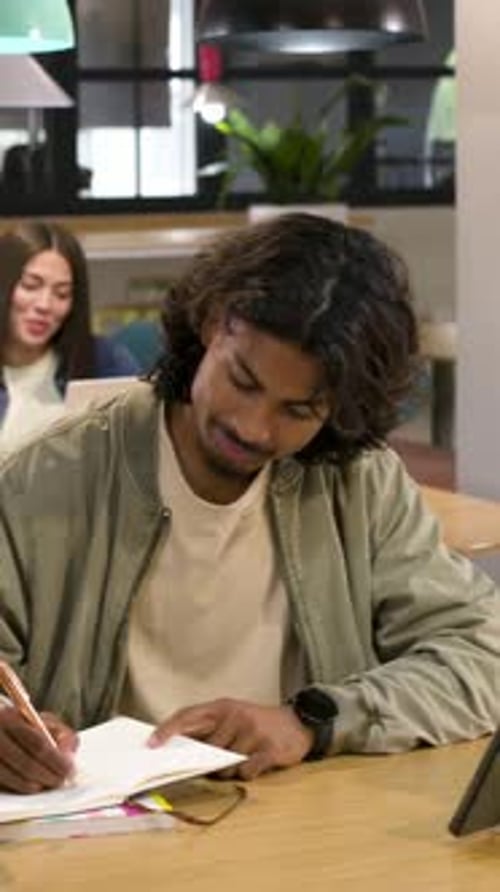 Vertical video: Writing male in green jacket at shared table with pen checking watch tapping tablet
