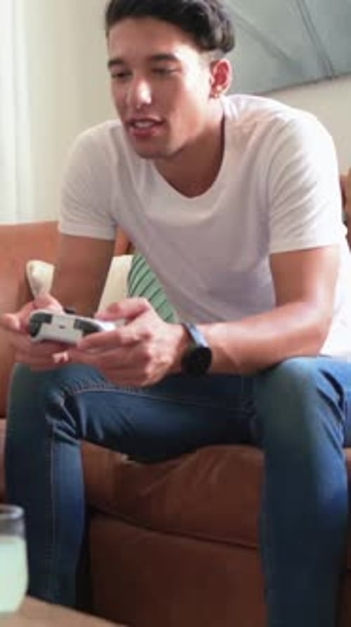 Vertical video: Leaning man in white tee pressing controller, reacting to game on couch
