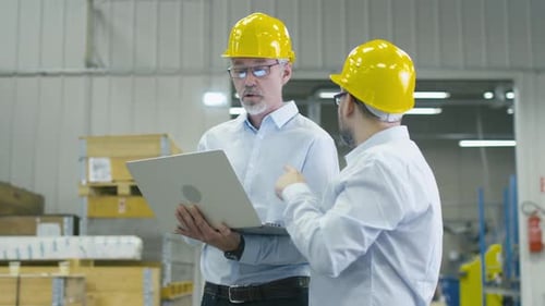 Two employees at logistics center warehouse are discussing work while holding a laptop computer.