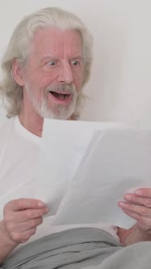 Senior Man Reads Paper In Bed and Reacts