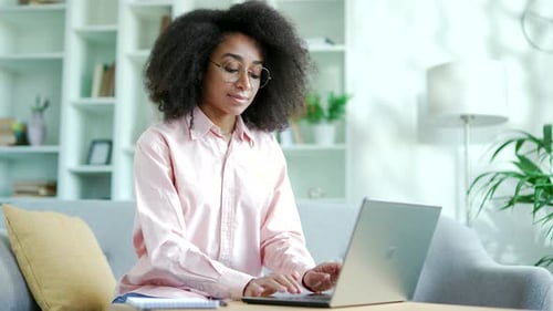 Young african american female freelancer works on laptop sitting on sofa at home office. Happy black