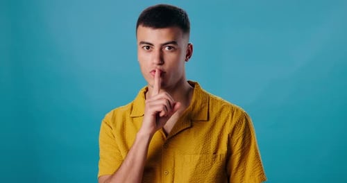 Face, finger on lip and man with silence, secret and confidential on blue studio background