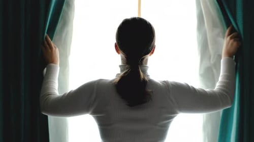 Woman stands at window and opens curtains rear view.
