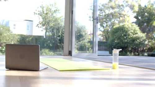 Close up of yoga mat, laptop and bottle with juice on floor at sunny home, slow motion